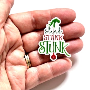 May include: A die-cut sticker featuring a green hand at the top, with the text "stink STANK STUNK" in black, red, and green lettering. A thin vertical line hangs from the hand to a small red Christmas ornament at the bottom.