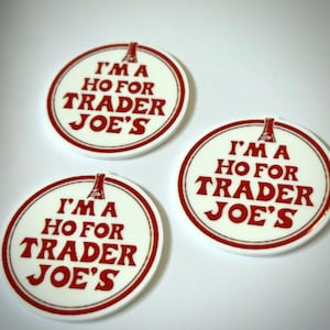 May include: Three white circular buttons with a red border and text that reads "I'M A HO FOR TRADER JOE'S" in red. The text is centered on the button, with a small illustration above the words.