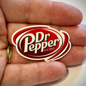 May include: A Dr Pepper logo sticker with a red and white design. The sticker features the Dr Pepper logo in white lettering on a red oval background, with the text "Est. 1885" below the logo.