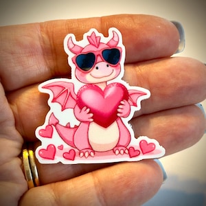 May include: A pink dragon sticker wearing sunglasses and holding a pink heart. The dragon has wings and is surrounded by small pink hearts. The sticker is a cute, whimsical design.