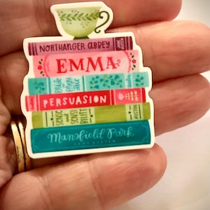 May include: A decorative pin featuring a stack of six colorful books titled Northanger Abbey, Emma, Pride and Prejudice, Persuasion, Sense and Sensibility, and Mansfield Park, topped with a small green and white floral teacup illustration.