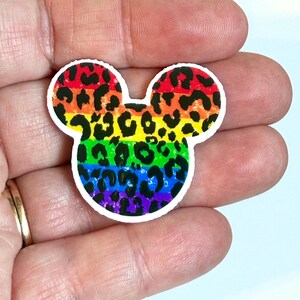 May include: A colorful sticker in the shape of a mouse head with rainbow stripes and leopard print. The sticker has a white border and is held in a hand. The colors include red, orange, yellow, green, blue, and purple.