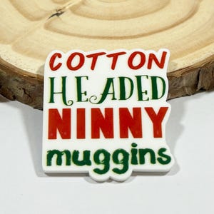 May include: A white rectangular acrylic pin with rounded edges featuring the text COTTON HEADED NINNY MUGGINS in alternating red and dark green lettering. The word COTTON is in red, HEADED is in green, NINNY is in red, and MUGGINS is in green.
