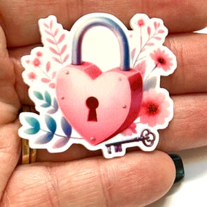 May include: A pink heart-shaped lock sticker with a silver handle and keyhole. The sticker is surrounded by pink flowers and green and blue leaves, and a decorative key. The sticker is held in a hand.