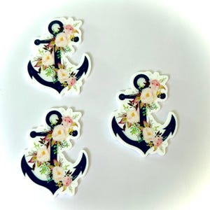 May include: Three navy blue anchor-shaped embellishments, each adorned with a floral arrangement of pink, white, and green flowers. The anchors have a white background and are suitable for crafts or nautical-themed projects.