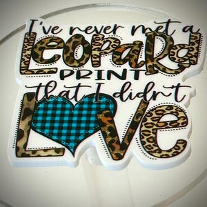 May include: A white background with a brown, black, and orange leopard print design. The design says "I've never met a leopard print that I didn't love" with a blue and white checkered heart in the middle.