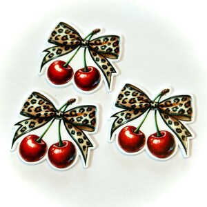 May include: Three stickers featuring a pair of glossy red cherries with green stems, topped with a leopard print bow. The stickers are on a white background. The cherries have a realistic appearance.