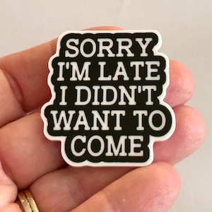 May include: A black and white rectangular pin with the text "SORRY I'M LATE I DIDN'T WANT TO COME". The pin has a white border and is held in a hand.