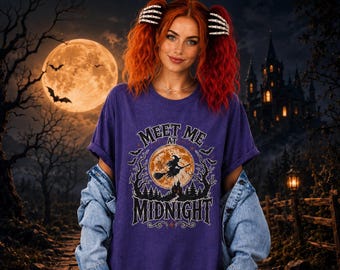 May include: A purple short-sleeve t-shirt featuring a graphic print of a witch flying on a broomstick in front of a full moon, surrounded by trees and bats, with the text "MEET ME AT MIDNIGHT" printed in a stylized, gothic-inspired font.