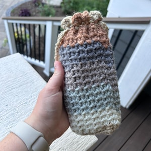 Peut inclure: Un manchon de bouteille crocheté avec un motif rayé multicolore dans des tons beige, marron, gris et bleu.