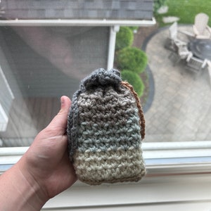 Könnte beinhalten: Eine gehäkelte Tasche mit Kordelzug. Die Tasche ist in einem gestreiften Muster aus braunem, grauem, blauem und cremefarbenem Garn gearbeitet.