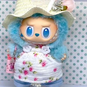 May include: A whimsical doll with blue fur, large blue eyes, and a toothy grin. It wears a straw hat with a butterfly accent, a floral dress, and denim pants. The doll is accessorized with white shoes and a pink water bottle.
