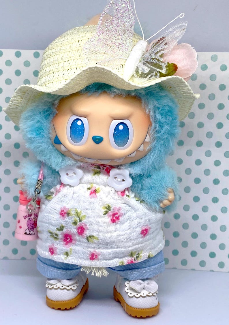 May include: A whimsical doll with blue fur, large blue eyes, and a toothy grin. It wears a straw hat with a butterfly accent, a floral dress, and denim pants. The doll is accessorized with white shoes and a pink water bottle.
