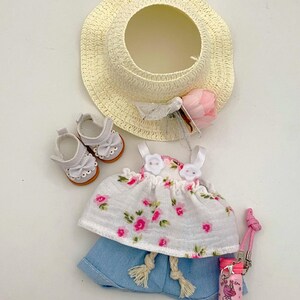May include: A doll-sized summer outfit set. Includes a straw sun hat, white floral top, denim shorts, white shoes, and a pink water bottle. The top features pink floral patterns. The hat has a pink flower and white ribbon.