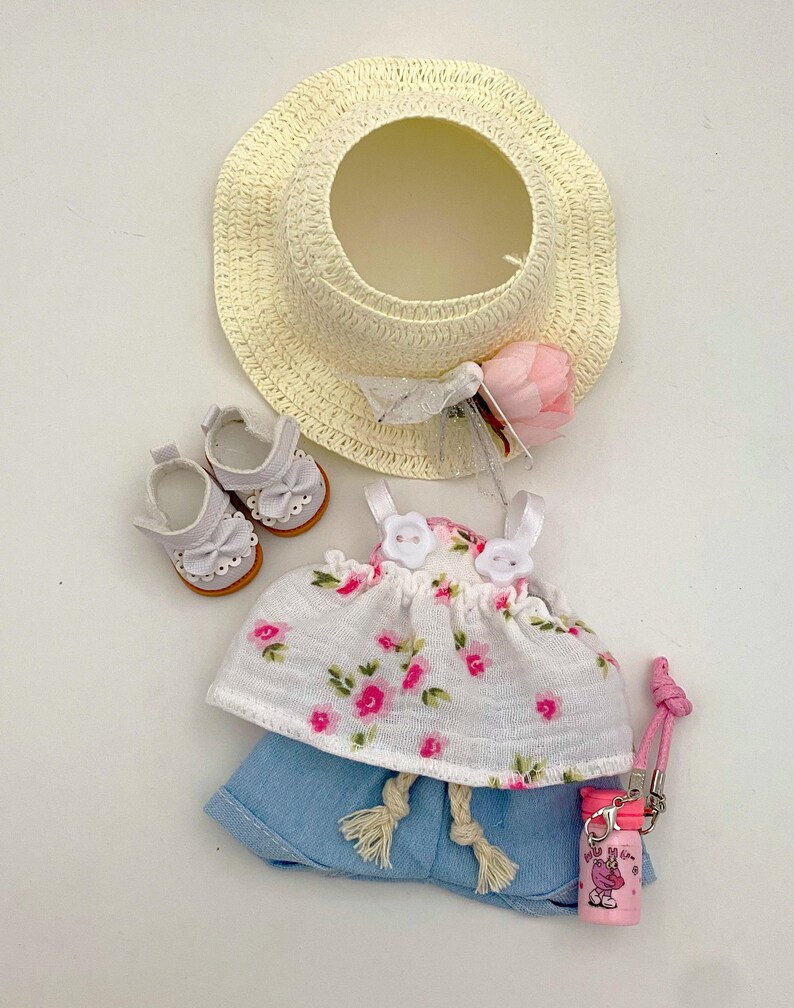 May include: A doll-sized summer outfit set. Includes a straw sun hat, white floral top, denim shorts, white shoes, and a pink water bottle. The top features pink floral patterns. The hat has a pink flower and white ribbon.