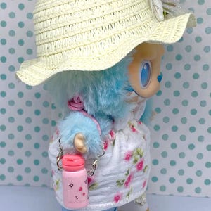 May include: A doll wearing a straw hat, a floral dress, and blue fur. The doll has a pink water bottle with a silver chain. The doll's outfit includes white shoes with a scalloped edge.