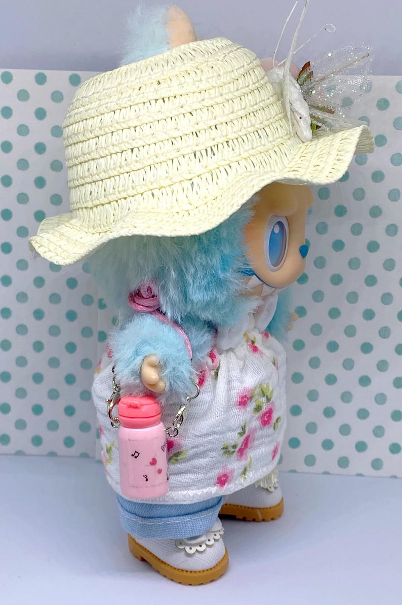 May include: A doll wearing a straw hat, a floral dress, and blue fur. The doll has a pink water bottle with a silver chain. The doll's outfit includes white shoes with a scalloped edge.