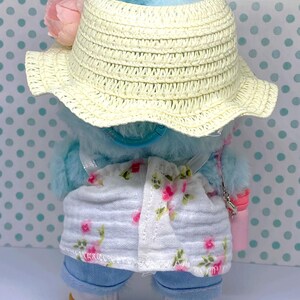 May include: A blue plush toy wearing a straw hat, floral print shorts, and blue jeans. The hat has a pink flower and blue feathers. The toy also has a small pink purse and white shoes with tan soles.