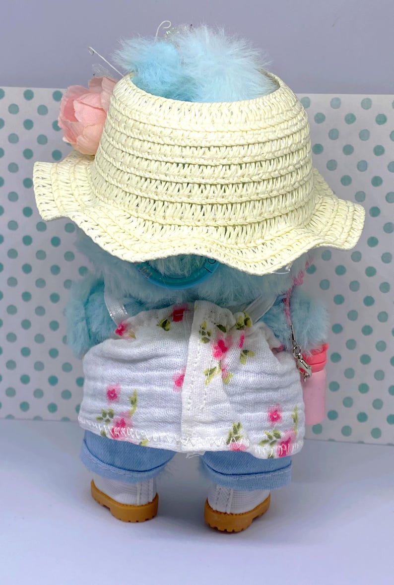 May include: A blue plush toy wearing a straw hat, floral print shorts, and blue jeans. The hat has a pink flower and blue feathers. The toy also has a small pink purse and white shoes with tan soles.