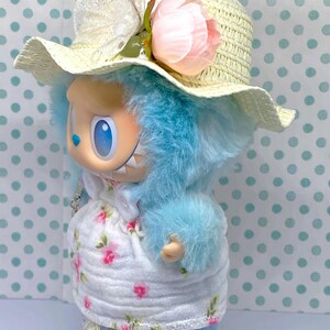 May include: A whimsical doll with blue fur, wearing a straw hat adorned with a pink flower and a white dress with a floral pattern. The doll is also wearing blue jeans and white shoes with decorative bows.