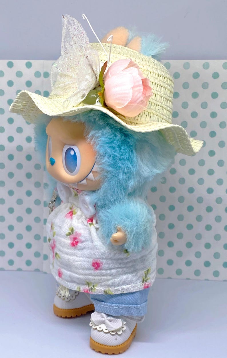 May include: A whimsical doll with blue fur, wearing a straw hat adorned with a pink flower and a white dress with a floral pattern. The doll is also wearing blue jeans and white shoes with decorative bows.