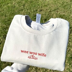 May include: Cream-colored sweatshirt with the words "wee woo wife" in red script, along with a red police car graphic. The sweatshirt is folded and resting on a bed of green grass.