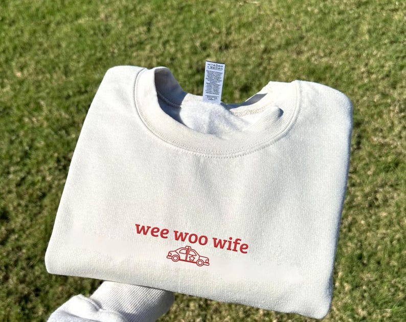 May include: Cream-colored sweatshirt with the words "wee woo wife" in red script, along with a red police car graphic. The sweatshirt is folded and resting on a bed of green grass.