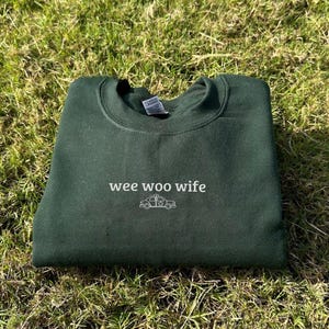 May include: A dark green sweatshirt with the words "wee woo wife" and a small ambulance graphic in white. The sweatshirt is folded and resting on a bed of green grass.