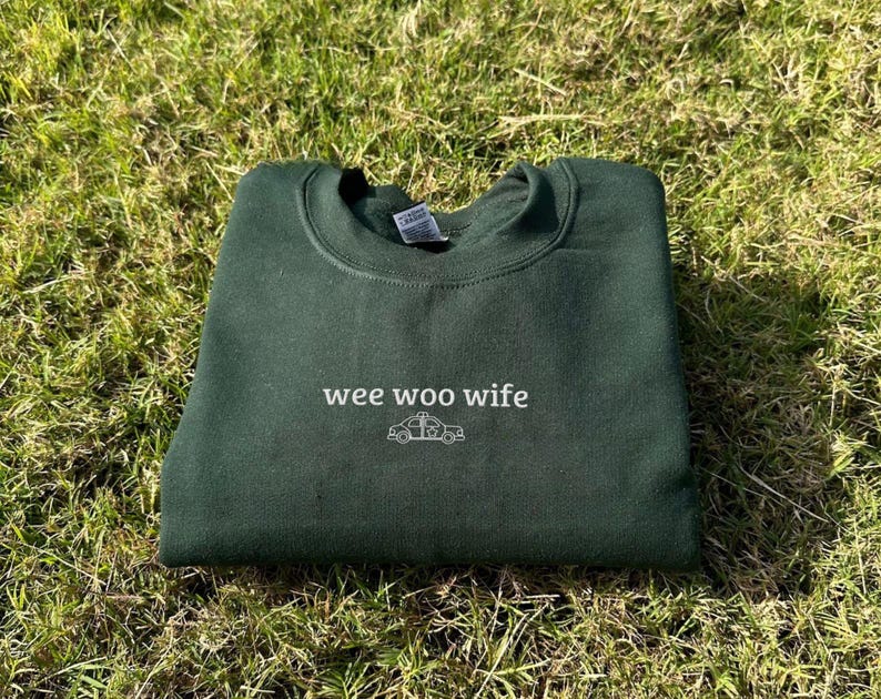 May include: A dark green sweatshirt with the words "wee woo wife" and a small ambulance graphic in white. The sweatshirt is folded and resting on a bed of green grass.