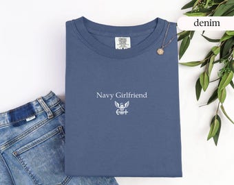 May include: A navy blue t-shirt with the words "Navy Girlfriend" and an emblem printed in white. The shirt is folded and placed next to a pair of denim jeans and a gold necklace. The word "denim" is written in a white oval.