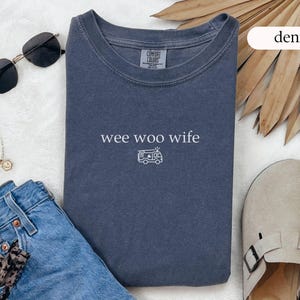 May include: A denim blue t-shirt with the words "wee woo wife" and a small ambulance graphic. The shirt is styled with sunglasses, a necklace, jeans, and sandals.