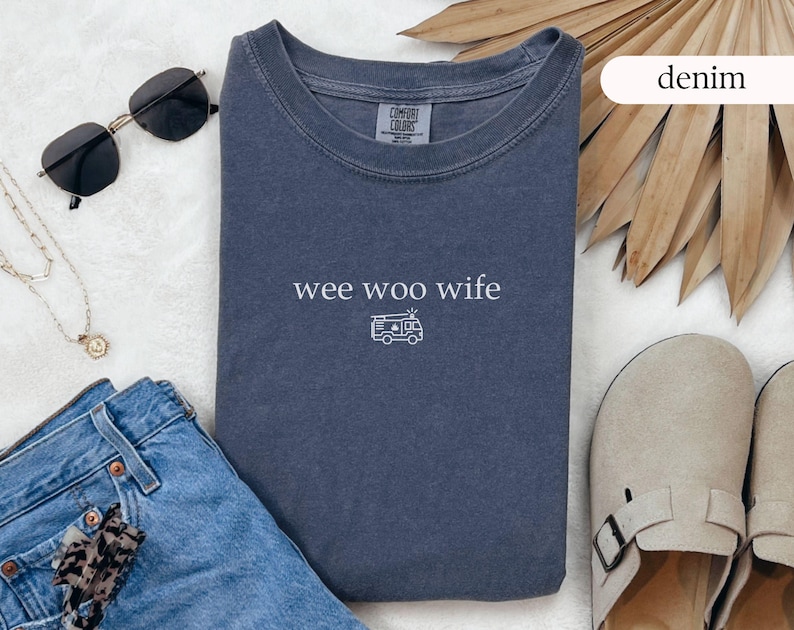 May include: A denim blue t-shirt with the words "wee woo wife" and a small ambulance graphic. The shirt is styled with sunglasses, a necklace, jeans, and sandals.