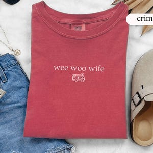 May include: Crimson-colored t-shirt with the text "wee woo wife" and a small graphic of a camper van. The shirt is displayed with blue jeans, sunglasses, and sandals.