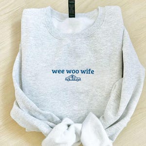 May include: A light gray sweatshirt with the words "wee woo wife" in blue script, along with a blue police car graphic. The sweatshirt is folded, with the sleeves and bottom hem visible.