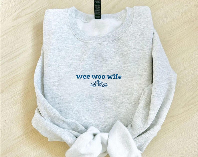 May include: A light gray sweatshirt with the words "wee woo wife" in blue script, along with a blue police car graphic. The sweatshirt is folded, with the sleeves and bottom hem visible.