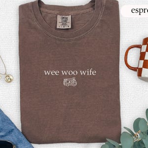 May include: Espresso-colored t-shirt with the words "wee woo wife" and a camper van graphic. The shirt is folded and has a crew neck. A mug with a checkered pattern and a gold necklace are also visible.