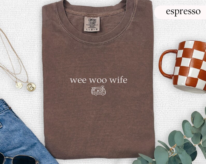May include: Espresso-colored t-shirt with the words "wee woo wife" and a camper van graphic. The shirt is folded and has a crew neck. A mug with a checkered pattern and a gold necklace are also visible.