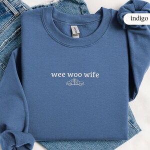 May include: Indigo blue sweatshirt with the text "wee woo wife" and a police car graphic. The sweatshirt is made of a soft material and is displayed on a pair of blue jeans.