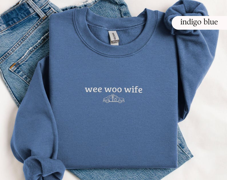 May include: Indigo blue sweatshirt with the text "wee woo wife" and a police car graphic. The sweatshirt is made of a soft material and is displayed on a pair of blue jeans.