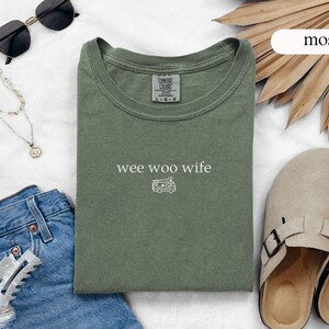 May include: A moss green t-shirt with the words "wee woo wife" and a small camper van graphic. The shirt is laid flat with sunglasses, a necklace, denim jeans, and sandals. The shirt is made of a soft, comfortable material.