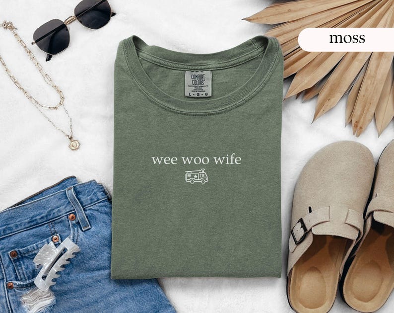 May include: A moss green t-shirt with the words "wee woo wife" and a small camper van graphic. The shirt is laid flat with sunglasses, a necklace, denim jeans, and sandals. The shirt is made of a soft, comfortable material.