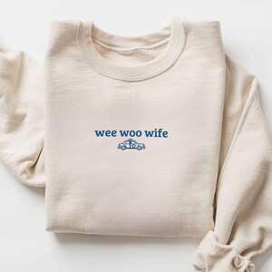 May include: Cream-colored sweatshirt with the text "wee woo wife" in blue, accompanied by a blue police car graphic. The crew neck sweatshirt is folded neatly, showcasing the design on the front.