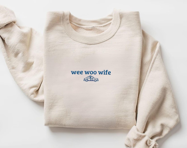 May include: Cream-colored sweatshirt with the text "wee woo wife" in blue, accompanied by a blue police car graphic. The crew neck sweatshirt is folded neatly, showcasing the design on the front.