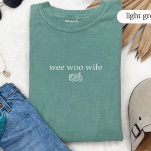 May include: Light green t-shirt with the text "wee woo wife" and a small camper graphic. The shirt is displayed with blue jeans, sunglasses, a necklace, and a pair of tan sandals.