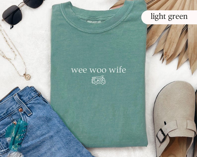 May include: Light green t-shirt with the text "wee woo wife" and a small camper graphic. The shirt is displayed with blue jeans, sunglasses, a necklace, and a pair of tan sandals.