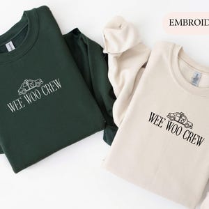May include: Two crewneck sweatshirts, one dark green and one cream-colored, are displayed. Both feature the embroidered text "WEE WOO CREW" with a police car graphic. The sweatshirts are made of a soft material and are suitable for casual wear.