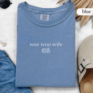 May include: A light blue t-shirt with the text "wee woo wife" and a small graphic of an ambulance. The shirt is folded and lies next to a pair of sunglasses, a necklace, and blue jeans.