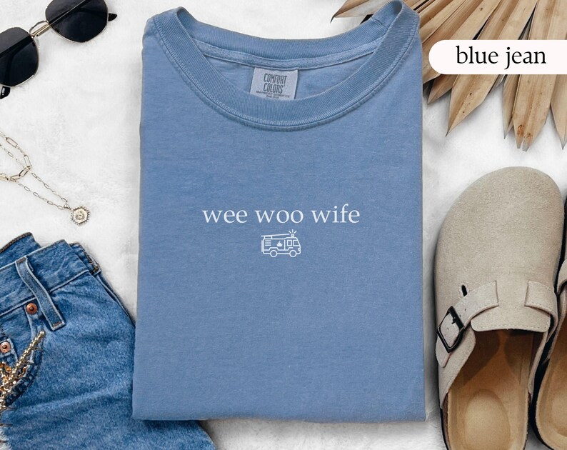 May include: A light blue t-shirt with the text "wee woo wife" and a small graphic of an ambulance. The shirt is folded and lies next to a pair of sunglasses, a necklace, and blue jeans.