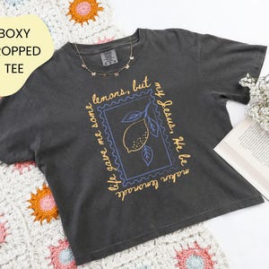 May include: Dark gray boxy cropped tee with a lemon graphic inside a blue postage stamp design. The text on the shirt reads "life gave me some lemons, but my Jesus, He be makin lemonade". A gold butterfly necklace is on the shirt.
