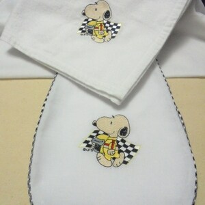 May include: Two white embroidered kitchen towels with a black and white checkered border. The towels feature a cartoon image of Snoopy dressed as a race car driver.